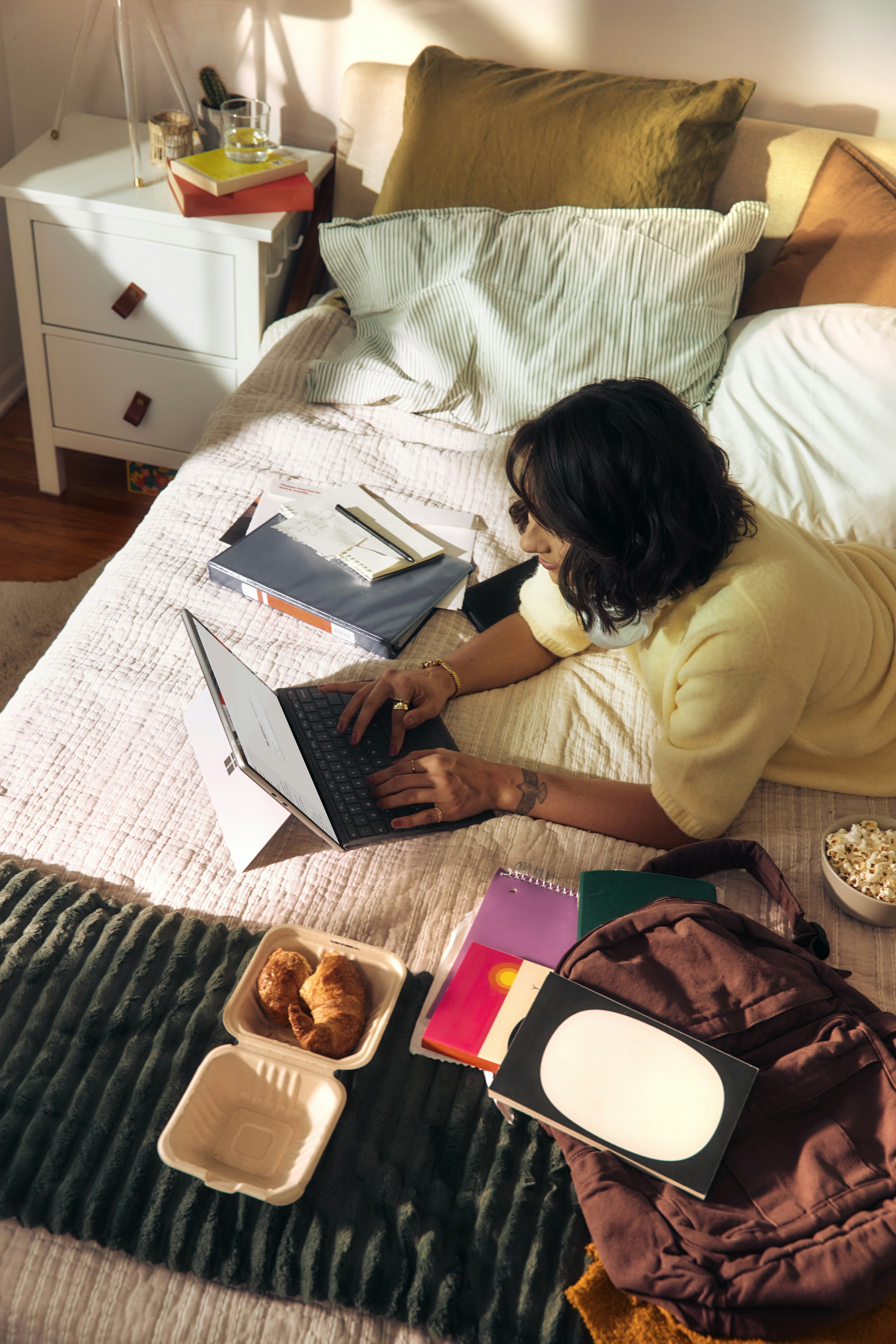A woman working on her laptop from her bed, surrounded by notebooks and papers